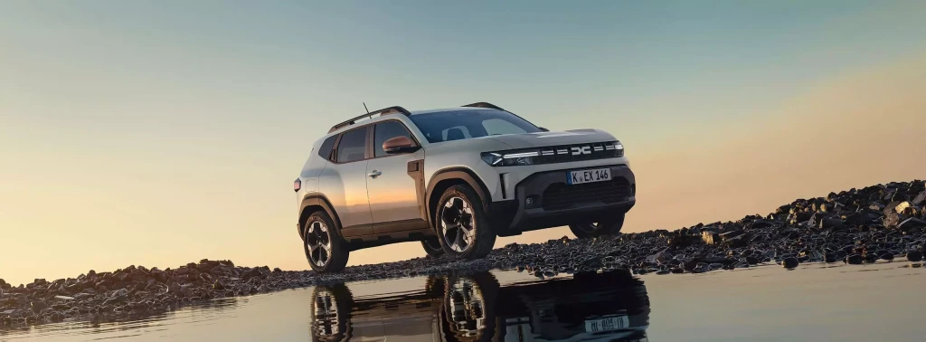 Silberner Dacia Duster SUV auf felsigem Seeufer bei Sonnenuntergang, gespiegelt im Wasser